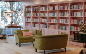 The interior of Enamoured Books in Remuera. Two plush green couches face each other in the shop floor, while the walls are lined with pink shelves full of brightly-coloured book spines.
