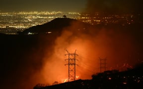 Flames and smoke are seen near power lines as the Palisades fire grows near the Mandeville Canyon neighborhood and Encino, California, on January 11, 2025. The Palisades Fire, the largest of the Los Angeles fires, spread toward previously untouched neighborhoods January 11, forcing new evacuations and dimming hopes that the disaster was coming under control. Across the city, at least 16 people have died as multiple fires have ripped through residential areas since January 7, razing thousands of homes in destruction that US President Joe Biden likened to a "war scene." (Photo by Patrick T. Fallon / AFP)