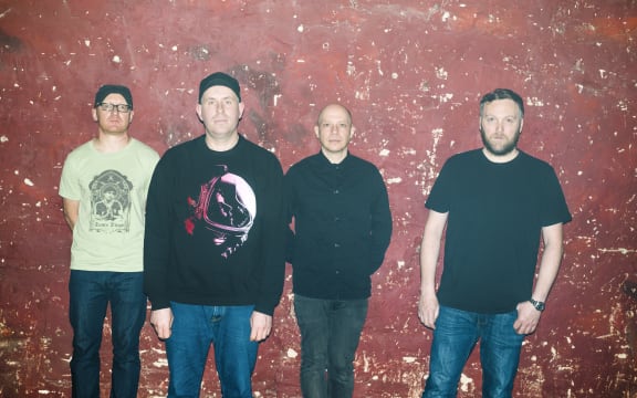 Members of Scottish band Mogwai are standing in a line against a red background.