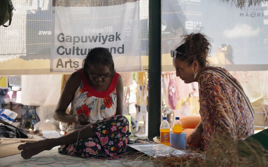Moana is sitting on the ground and talking with an indigenous woman who is busy weaving. They are at a busy marketplace.