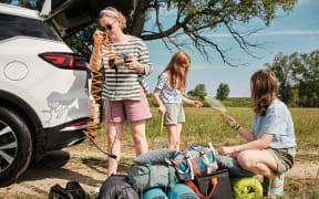 A family unpack their car with road trip items including bags and tennis rackets.