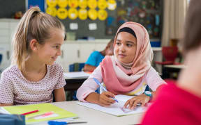 Young arab girl with hijab doing exercise with her bestfriend at international school. Asian muslim school girl sitting near her classmate during lesson. Multiethnic elementary students in classroom.