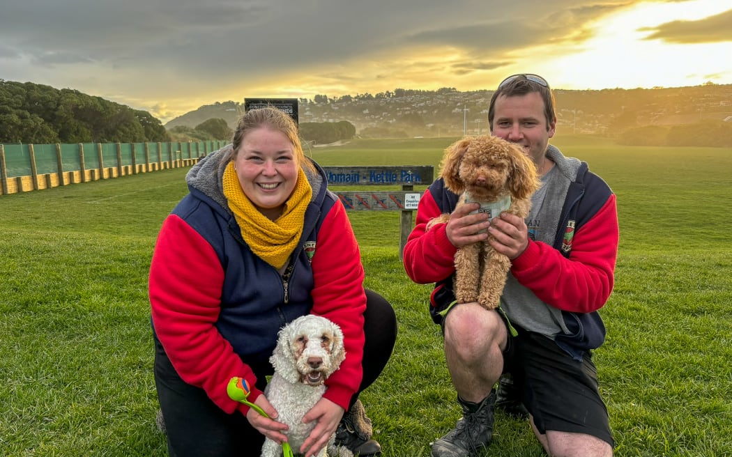 A man and a woman crouch down in a grassy field with hills far in the background and a golden sunset. They are both wearing red and grey hoodies. The man has a small light brown dog on his knee, and the woman has her hands cradling a white dog sitting on the ground. Both dogs have curly coats suggestive of a poodle cross breed.