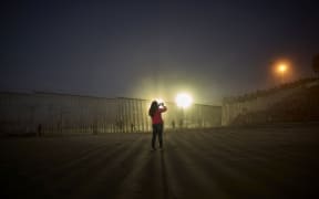 A woman records with her phone, as floodlights from the United States light up the border wall, topped with razor wire, along the beach in Tijuana, Mexico.