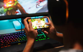 Teenage gamer boy playing video games on smartphone and computer in dark room wearing headphones and using backlit colorful keyboard