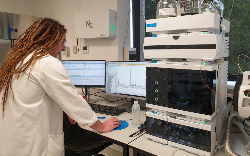 A man with long dreadlocks wearing a white lab coat looks at a computer monitor displaying a graph with sharp peaks on it. Next to him is a large machine.