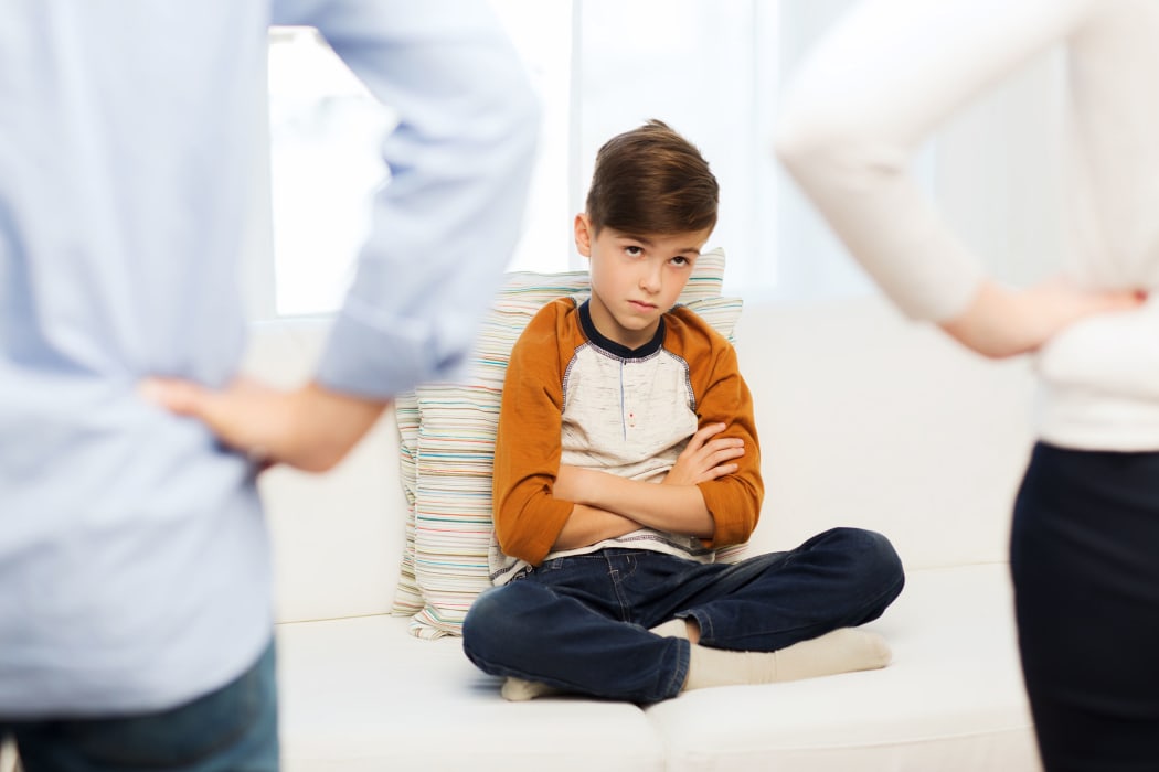 Small boy looking guilty sits on the couch faced by two parents with their hands on their hips