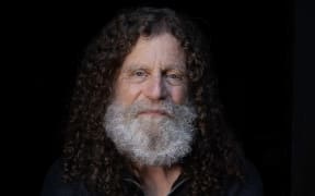 Robert Sapolsky has long brown hair and a white beard and sits on a stool holding what appears to be a human skull.
