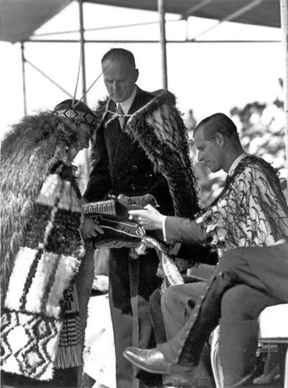The Duke of Edinburgh receives a gift during the Māori reception at Rotorua while Minister of Māori Affairs E.B. Corbett looks on, 2 January 1954.