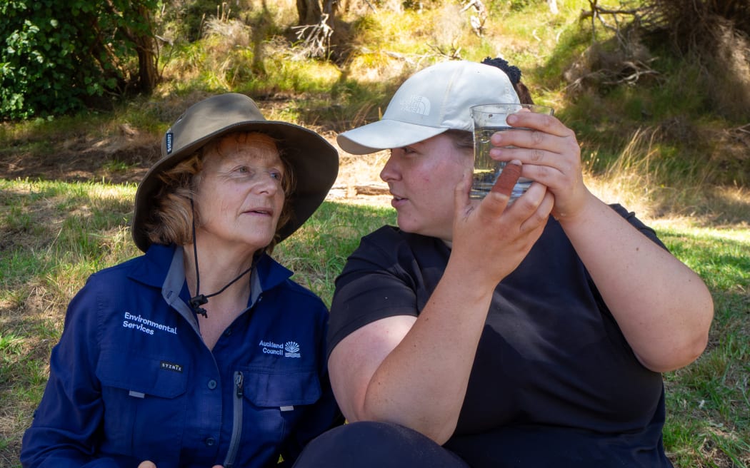 Two women sit on a grassy verge. The one on the right is wearing a white cap and black t-shirt, and is holding up a glass jar with her hands, while the other woman is looking intently at the jar. The woman on the left is wearing a wide-brimmed hat and navy blue Auckland Council-branded collared shirt.