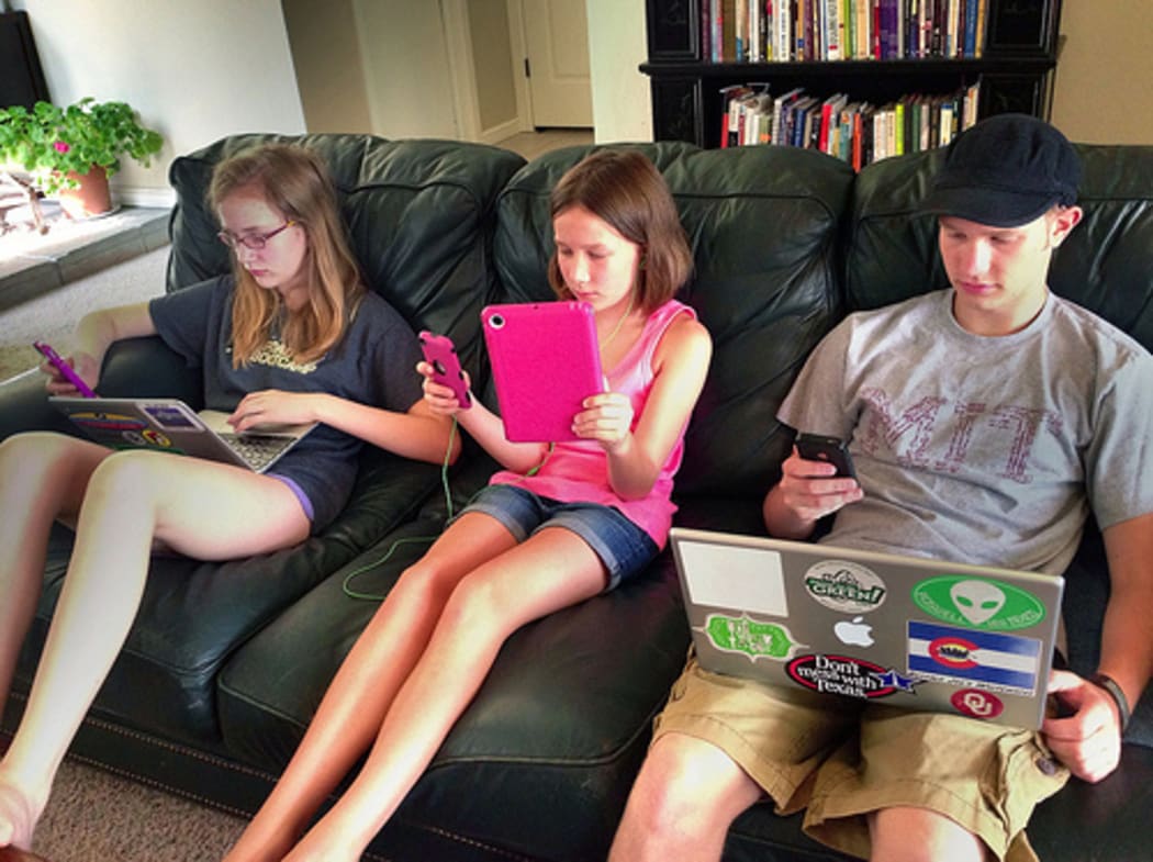 A photo of three children on a couch, all using multiple devices