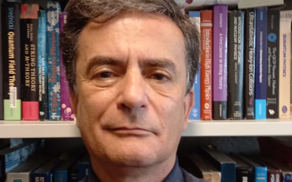 Dr David Krofcheck stands in front of a colourful bookshelf. He is wearing a dark collared shirt with a black tie. He is looking straight at the camera.