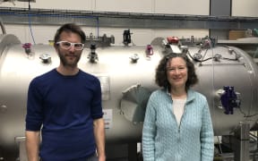 A man and a woman stand in front of a large metallic cylinder with various valves in a lab.