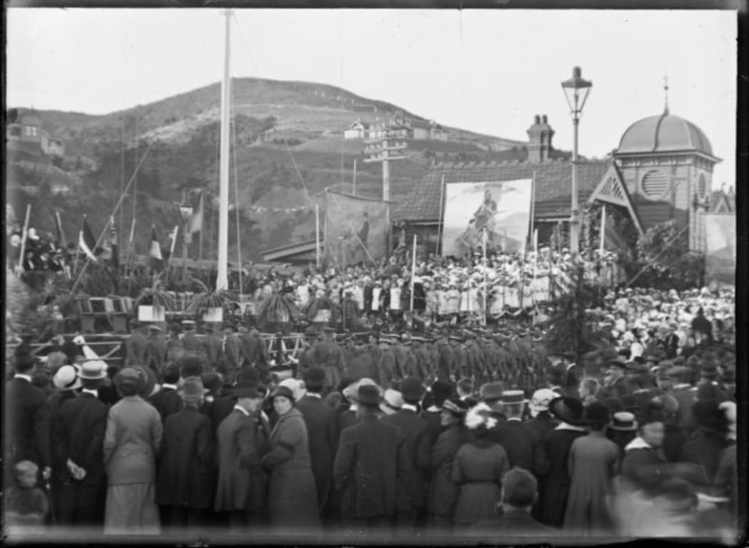 Anzac day commemoration at Petone, on 25 April 1916. Shows a crowd, flagpole and banners next to the Petone Railway Station. Photograph taken by Albert Percy Godber.
