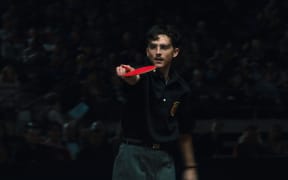 Timothee Chalamet, a young man with a moustache and glasses, holds a red ping-pong racquet as he points a finger.