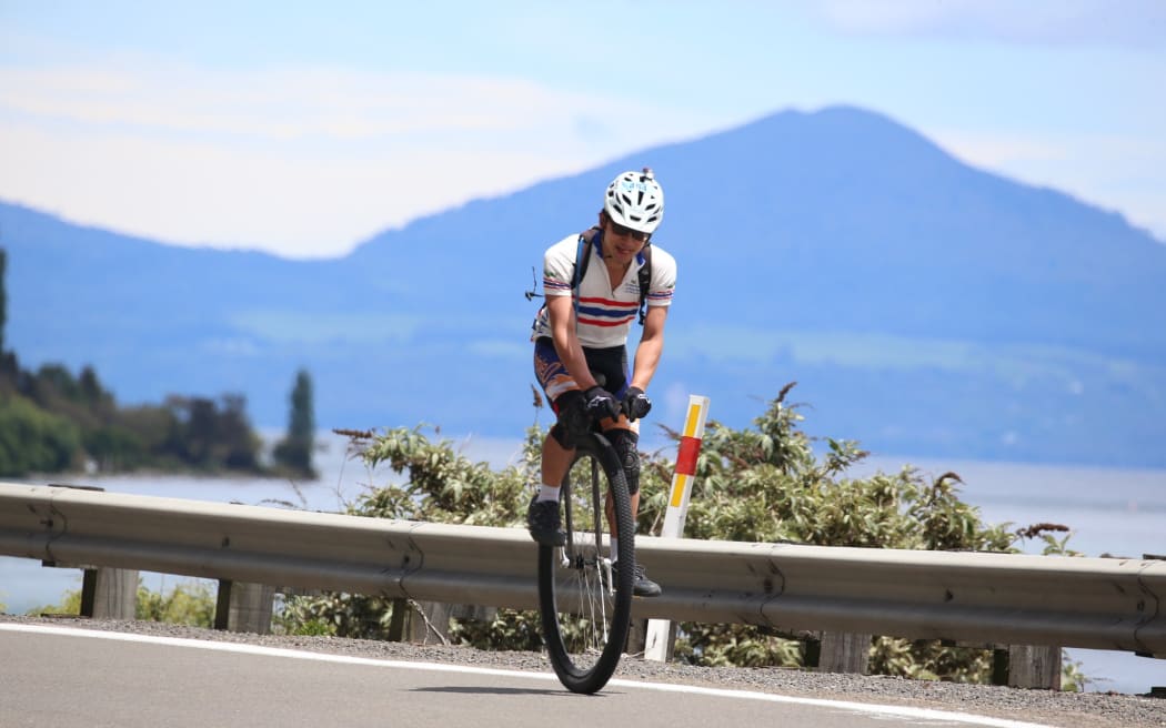 Ken rides a unicycle on a road, with a lake in the distance.
