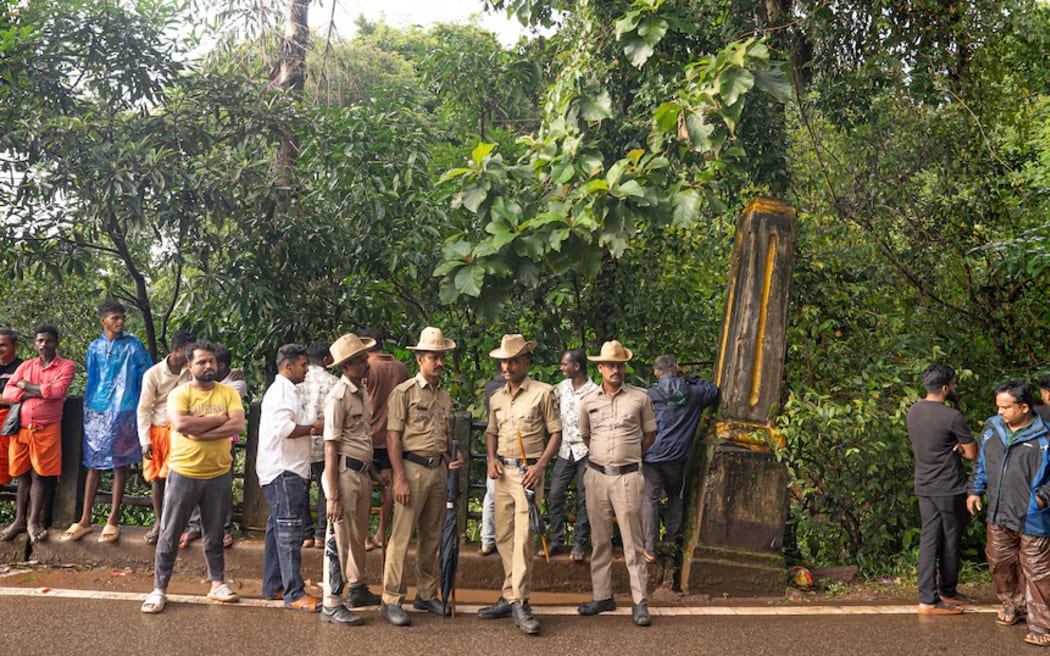 Indian police officers in tan clothing and hats standing on a roadway next to a line of men, a concrete monolith and forestry.