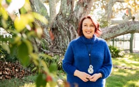 neuroscientist Christine Ozolins wearing a blue sweater stands in front of a large tree with some leave out of focus in the foreground