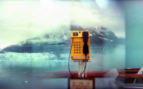 Glacier Bay, Alaska, June, 2022. (L): A glacier in Glacier Bay, Alaska. (C): An emergency phone on the deck of a cruise ship parked at the glacier. (R): A cruise passenger stands on the balcony of their cabin on a cruise ship. The summer cruising season is typically 4 months long, but is now getting stretched to 7 months, with the combination of global warming and larger ships being able to navigate through The Last Frontier