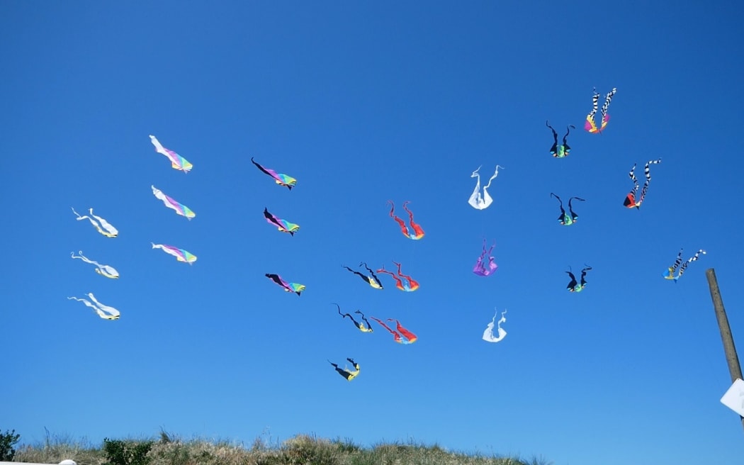 A colourful assortment of flow tail kites fly in formation against a bright blue sky.