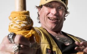 Man in yellow cap  saying "Tribal Huk," and yellow leather jerkin,  holding out hand grasping a big hammer. Hand has 3 big rings and man is staring.