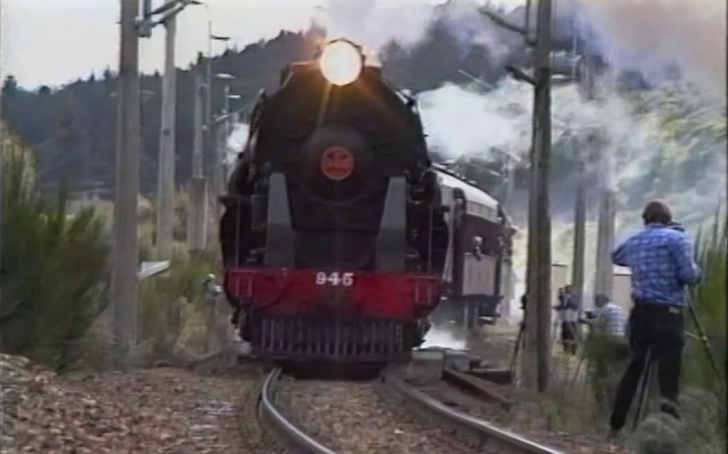 A steam train is headed towards the camera, a man in a blue shirt is beside the rail track filming the train go by on a camera.
