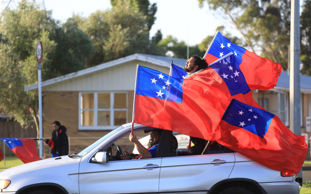 A photo of Toa Samoa supporters in Auckland, 20 November 2022. A car passes with one suppporter standing up through the sunroof. Four samoan flags are flying from the car. Toa Samoa were defeated by Australia 30-10 in the  Rugby League World Cup final in Manchester.