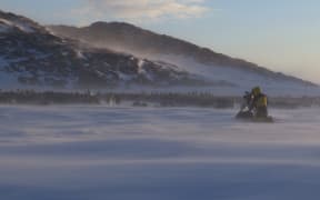 A photo of the Antarctic tundra. Frederique kneels by a camera tripod, filming a huge gathering of emperor penguins huddled in the misty morning light.