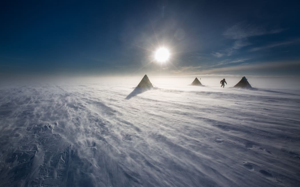 Three pyramidal tents on a flat expanse of white ice beneath a harsh sun and darkened sky. A lone figure walks between two tents.