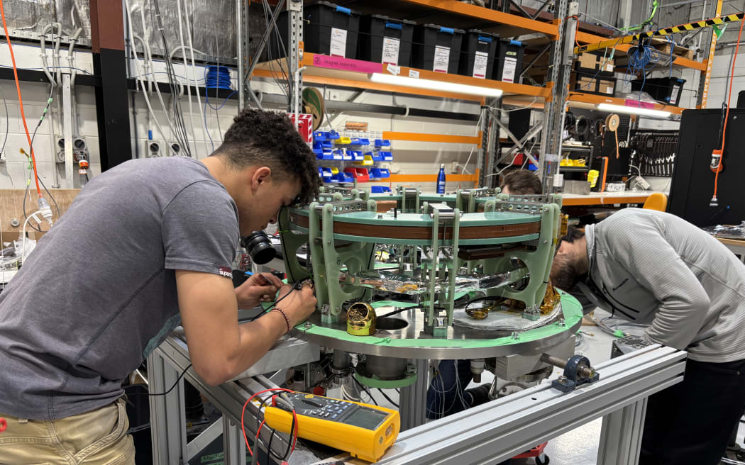 Two people working on a complex circular metal object in a lab.