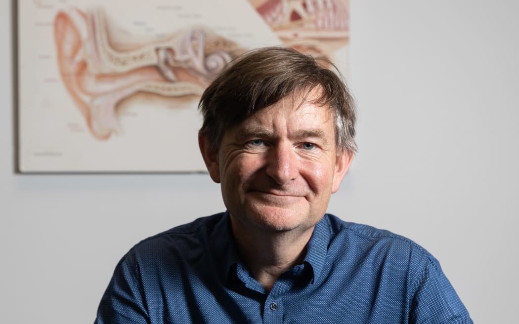 A portrait of Grant. He is in front of a diagram of the anatomy of the ear. He has a wry smile and is wearing a blue shirt.