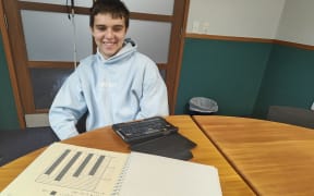 Toby Ireland smiles as he sits in front of a desk with a booklet showing a graph and his BrailleSense device.