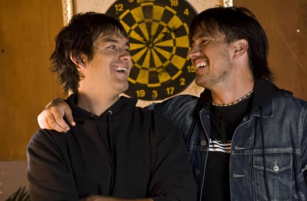 Two dark-haired young men smile widely at each other with their arms around each other.