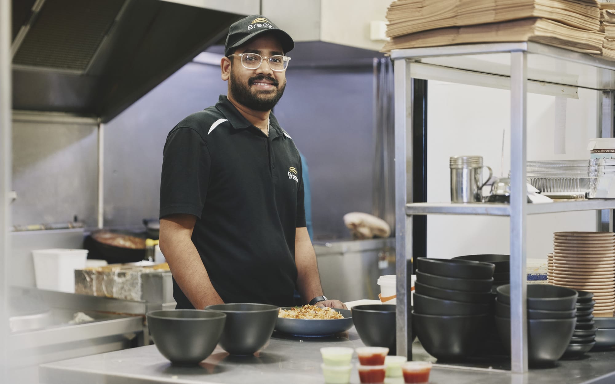 Noor Mohammed Shaikh, wearing a black polo shirt and black cap, poses in the kitchen of his Hamilton restaurant, Breeze.
