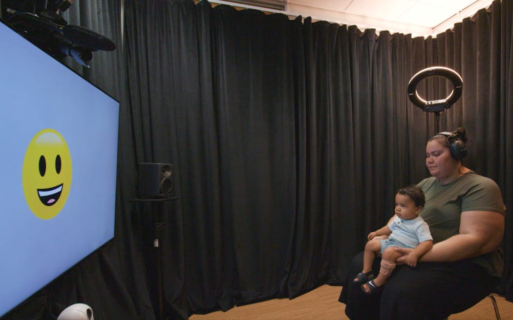 A woman sits on a chair with a baby on her lap underneath a ring light. The woman is wearing headphones and the baby is looking at a large screen with a smiley face emoji on it.