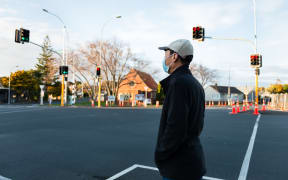 Man wearing a facemask waiting at the intersection for the green pedestrian light. Roadwork traffic cones along the street.