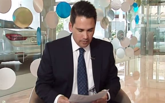 Simon Bridges looks at Newshub;s leaked list of his travel expenses during Monday's exclusive report  - with his crown car in the background.