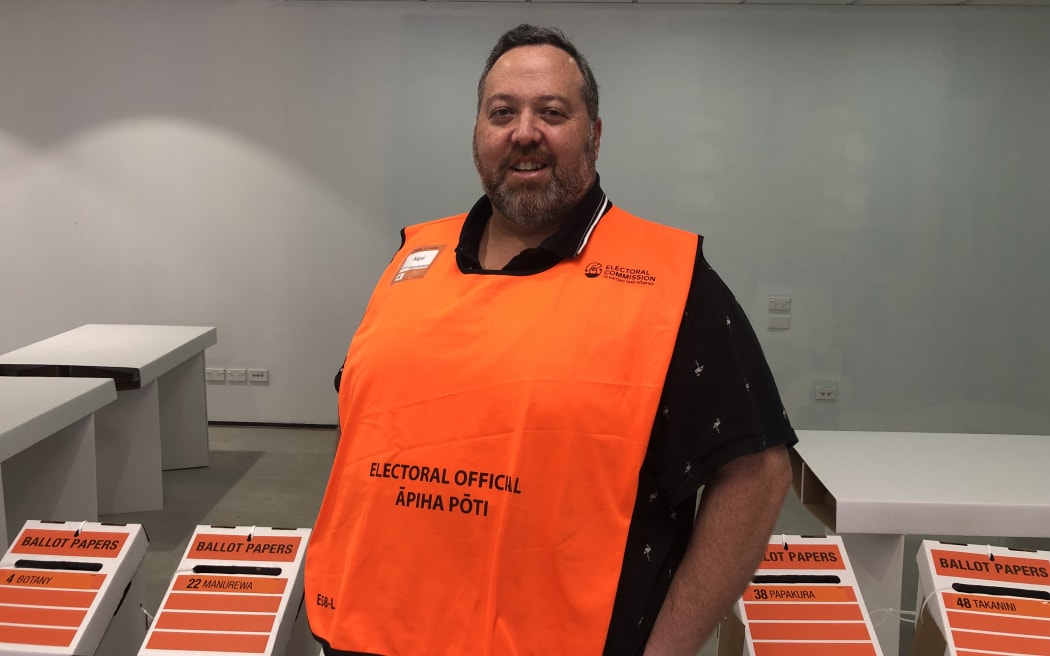 Nigel smiles at the camera. He is wearing a bright orange vest that reads "ELECTORAL OFFICIAL - ĀPIHA PŌTI". To his left and right are ballot boxes for people to drop their votes into.
