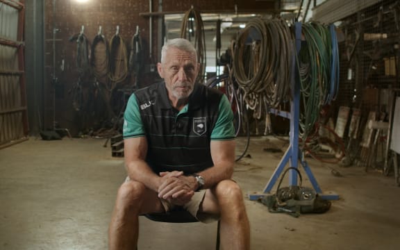 Mark Graham sits on a chair in a warehouse, looking intently at his interviewer to the right of the camera. He wears a green tshirt and a black vest.