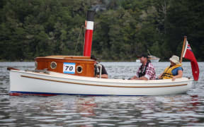 A vintage tugboat with a red chimney. Three people sit in the boat as it glides along a glassy lake, with trees on the shore in the background.