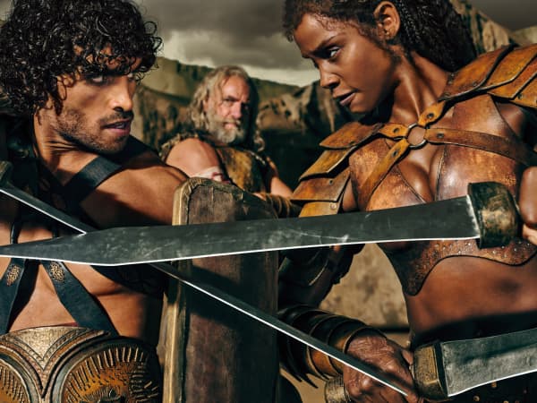 A man in woman in gladiator costumes face each other as they cross swords.