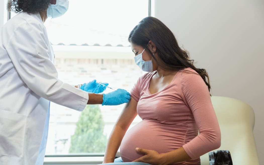 Wearing protective masks because of COVID-19, a mid adult pregnant woman watches as the female technician administers a booster shot.