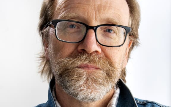 George Saunders wears a greying beard, dark-rimmed glasses and a denim acket.
