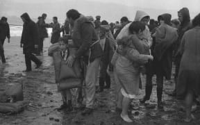 Clarence O'Neill (centre, wearing life-jacket) on Seatoun beach after being rescued from the Wahine shipwreck. The man with the boy who is wearing a life-jacket is John McDougall of Martinborough. Photographed 10 April 1968 by an Evening Post staff photographer.
