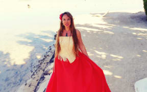 Toni Huata stands on a white sand beach, wearing pounamu and a red dress with a red frangipani in her hair.