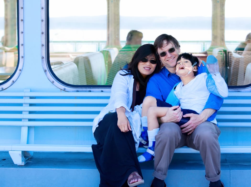 A photo of a smiling multiracial, interracial couple holding disabled son on ferry boat deck. Their child has cerebral palsy.
