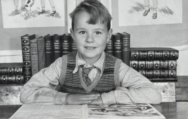 Tom, a young boy with a gentle smile wearing a tie and woollen vest, looks at the camera with a book open in front of him.
