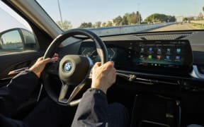 A driver's hand operates the steering wheel and reaches toward the curved digital infotainment screen of a BMW electric vehicle (EV) while driving on a street in Bari, Italy, on November 22, 2025. The wide screen displays the main menu, with icons for various apps and settings in Italian, such as 'Tutti' (All) and 'Infotainment', demonstrating the vehicle's integrated digital interface. (Photo by Matteo Della Torre/NurPhoto) (Photo by Matteo Della Torre / NurPhoto via AFP)