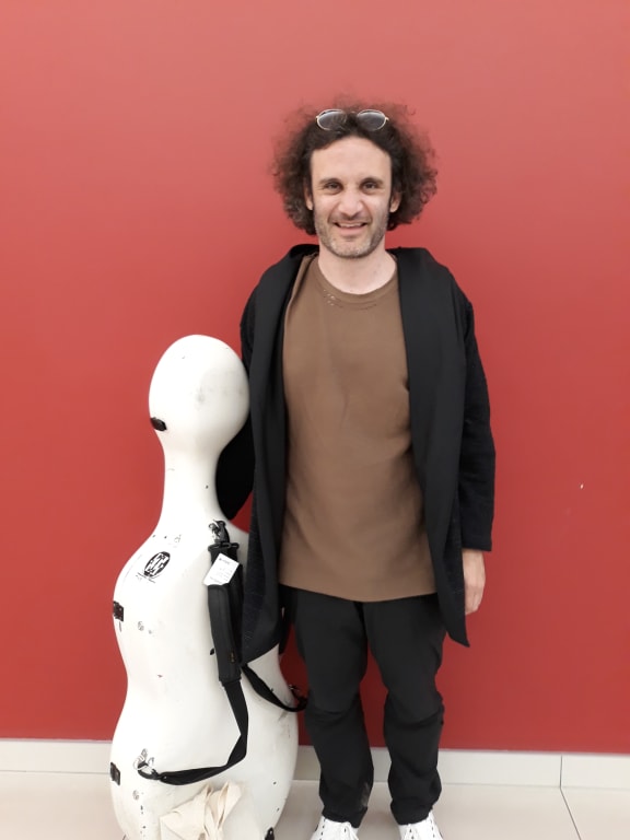 Cellist Nicolas Altstaedt stands beside his white cello case, against a red wall.