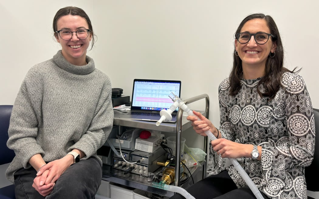 Two women with long dark hair and glasses smile and sit next to a laptop on a metal trolley in front of a white wall. On the tray underneath the laptop is a complex machine with a long tube, that the woman on the right is holding in her hands.